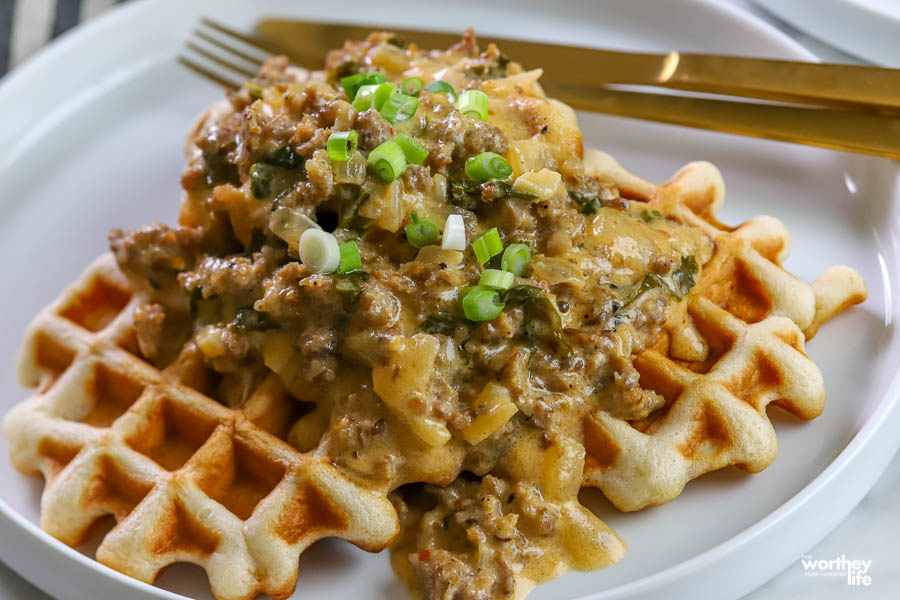 waffles with sausage gravy piled on top plated on a white plate with gold flatware